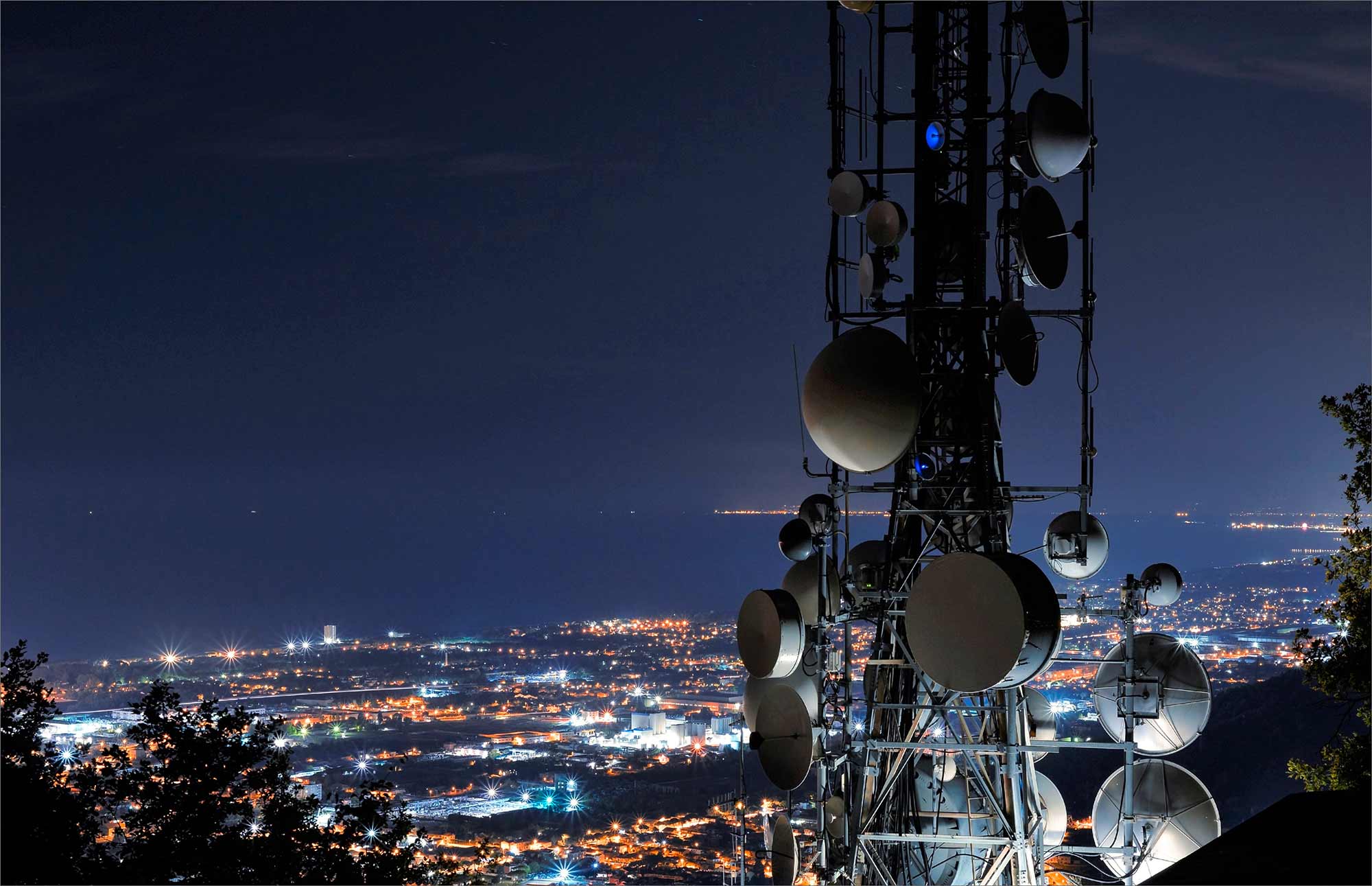 Tour de télécommunication avec antennes et paraboles surplombant une ville illuminée la nuit